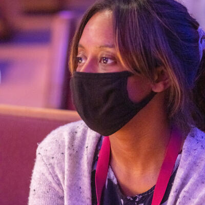 Sitting the the maroon pews of the Keene Church, a woman sits, wearing a black mask, and looks forward at the speaker.