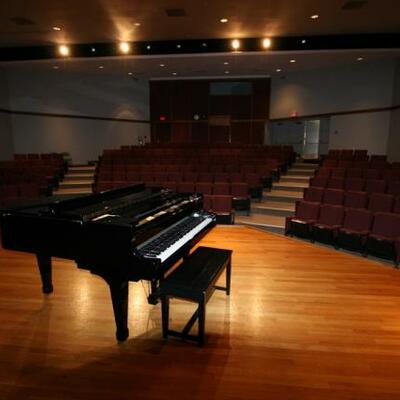 A black, shiny piano sits on a wooden stage in front of maroon chairs installed in the auditorium