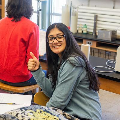 A student smiles and gives a big thumbs up as she prepares for class with her notes and backpack in front of her