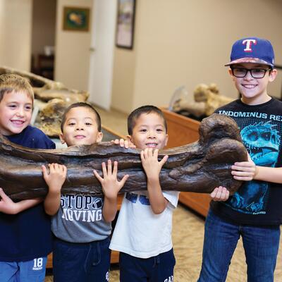 Four young boys smile big as they successfully carry a dinosaur bone that is much bigger than them