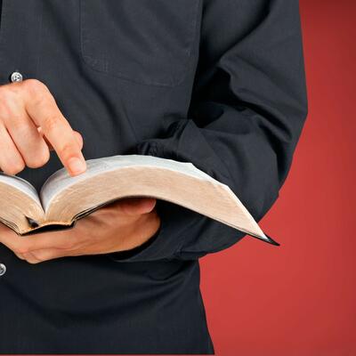 A man dressed in a black button-up holds a bible with one hand and points to a specific verse
