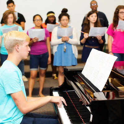 A pianist reads sheet music while playing for the choir that is practicing behind him