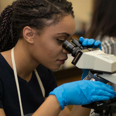 A student wearing blue gloves looks down at microscope as she inspects bacteria