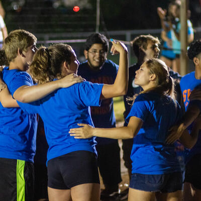 Large group of students in matching blue shirts gather in a team huddle in an outdoor volleyball court filled with mud