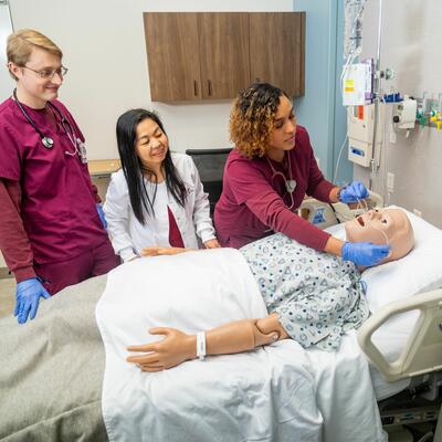 A nursing student, dressed in maroon scrubs, places oxygen tubes in a fake patients nose while a peer and professor stand by and watch