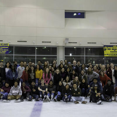 A large group of students smile big as they huddle for a group photo on the ice of the ice skating rink