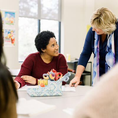 A professor leans over as she talks with a student sitting at a round school table