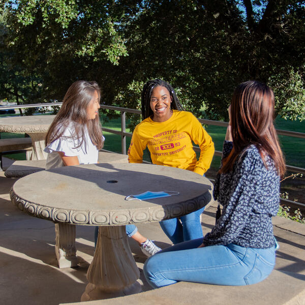Three female students sit on the patio in the sunny weather and laugh