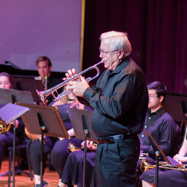 Professor with grey hair and glasses, playing goldeen trumpet at a university recital. 