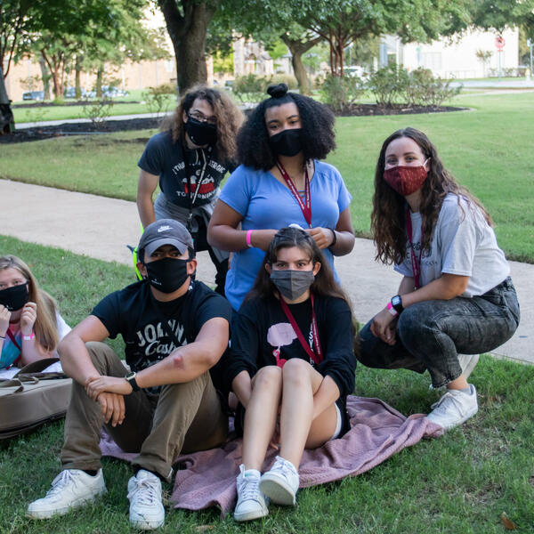 Sitting on blankets outside, six friends gather close and smile big under their face masks