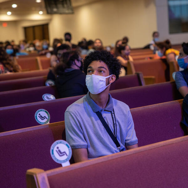 Students wearing face mask sit distanced in maroon pews in the church and look up