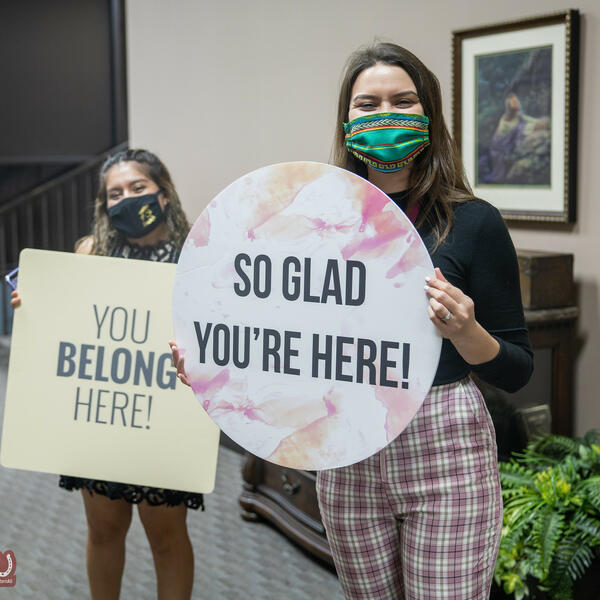 Two female students wearing masks hold up signs with welcome greetings at vespers