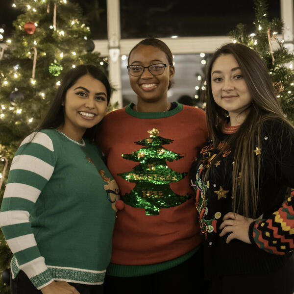 Three young ladies smiling, wearing red and greenn Christmas sweaters, standing together in front of two decorated Christmas trees with their arms around eachother 