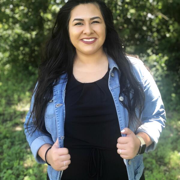 A female student with dark hair smiles as she wears a black jumper and jean jacket