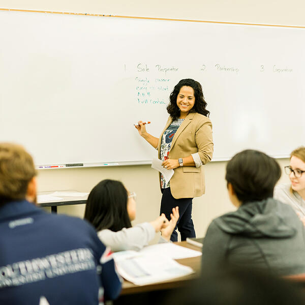 A business professor smiles and turns from writing on her board as she listens to a students question