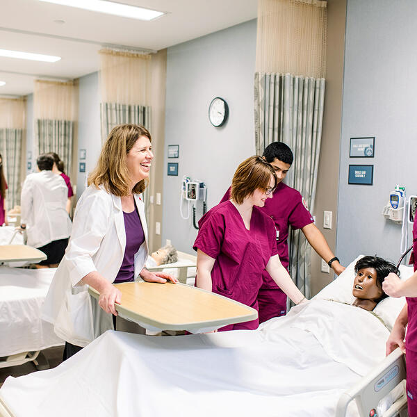 Three nursing students dressed in maroon scrubs look down at their patient and their professor watches over and smiles