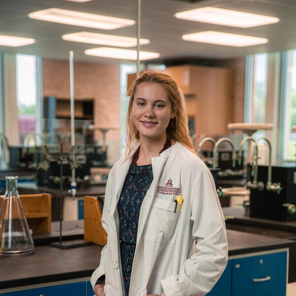 Kelsey Johnson poses in the lab wearing a lab coat that reads "Southwestern Adventist University Chemistry" with her hands in her pockets