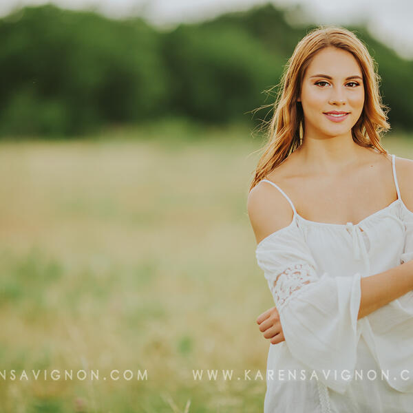 Female student gently crosses her arms as she wears a white dress and stands outside in a grassy field