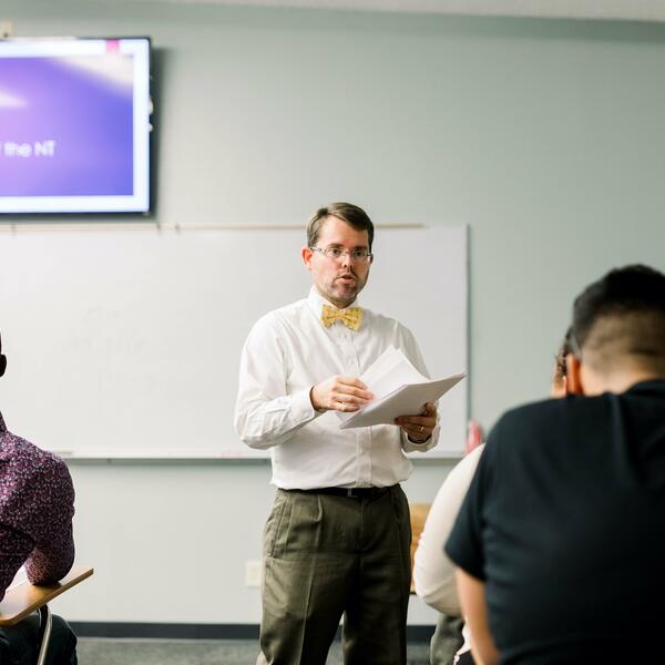 Dr. Campbell, dressed in a white buttoned shirt and yellow bow-tie, stands in front of his class and holds papers as he begins to hand them out to his students