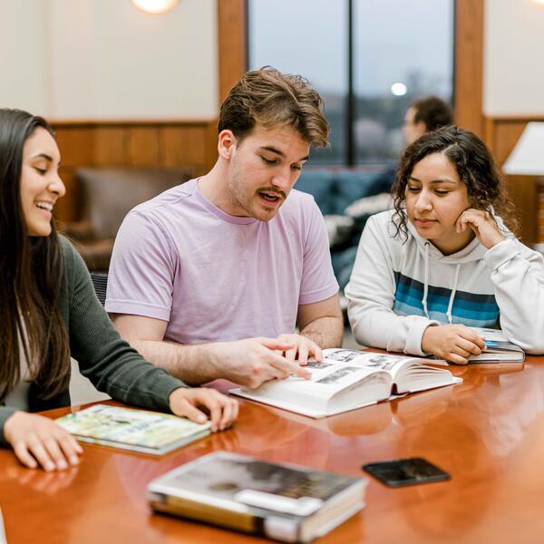 Three students look at a book as they sit at a rectangular table in the pechero student lounge