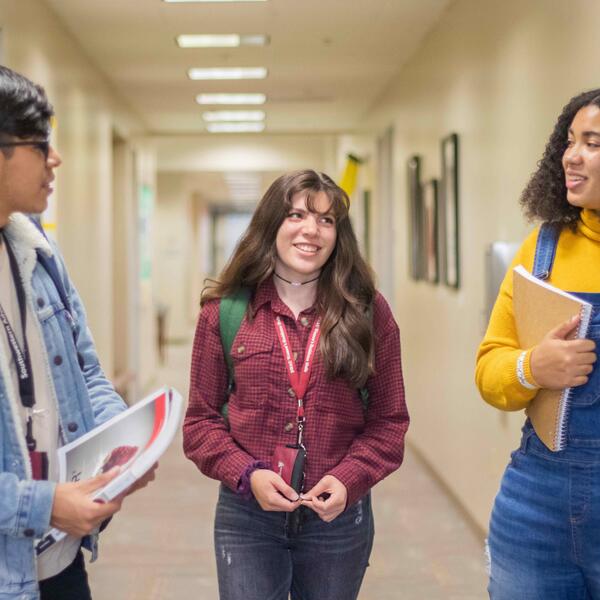 Three students stand in a hallway with books, talking