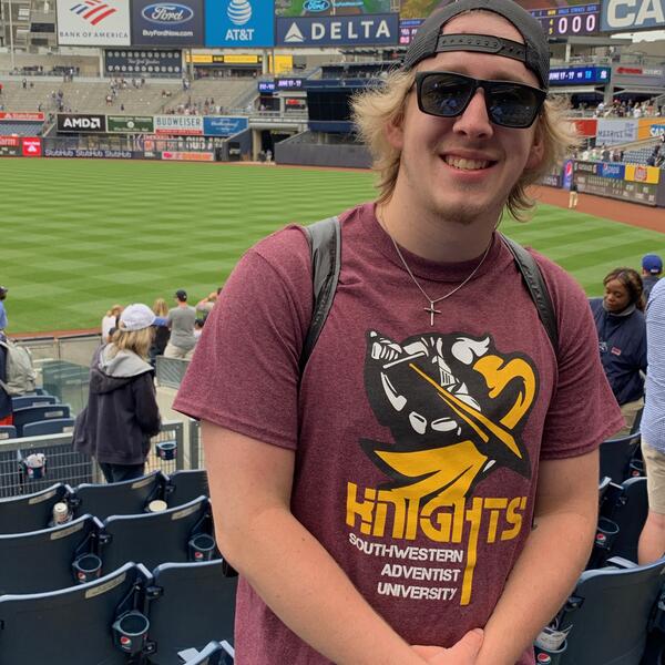 Male student smiles as he stands in the bleachers of the Yankee stadium wearing a SWAU Knights shirt