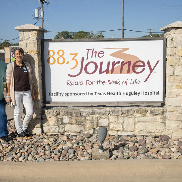 Mike and Wanda Agee stand to the left of the radio station sign that reads "88.3 The Journey, Radio for the Walk of Life." Danae stands to the right of the sign.