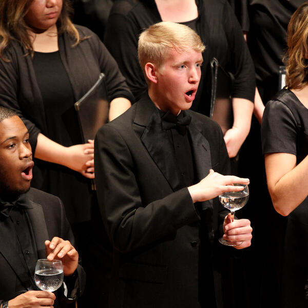 A group of singers, wearing all black, sing as the touch the rim of glasses filled with water