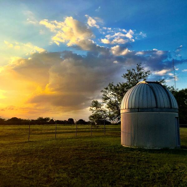 The obervatory, in a silver color, sits on grass with a fence surrounding it