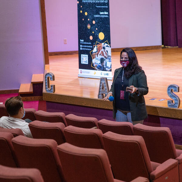 Mrs. Patterson stands, holding a mic, in front of an auditorium with two students in the seats in front of her