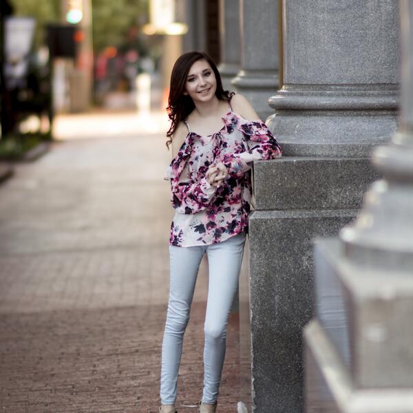 Student, wearing a white and pink blouse with light blue jeans, leans on a pillar outside as she smiles 