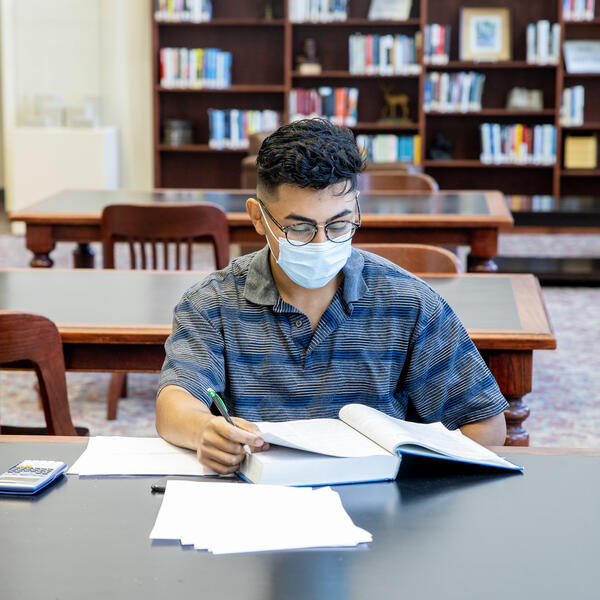 Student wearing a mask sits at a table in the library in front of an open textbook