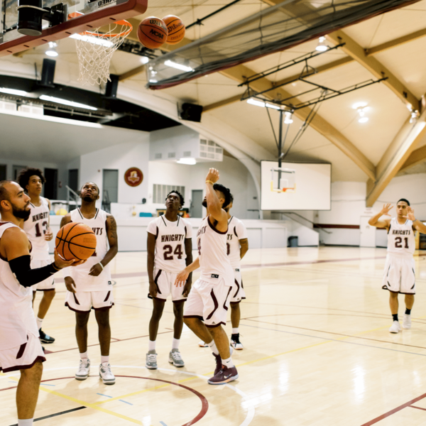 A group of male basketball players gather in the gymnasium wearing their uniforms as thye throw balls into the hoop and look around