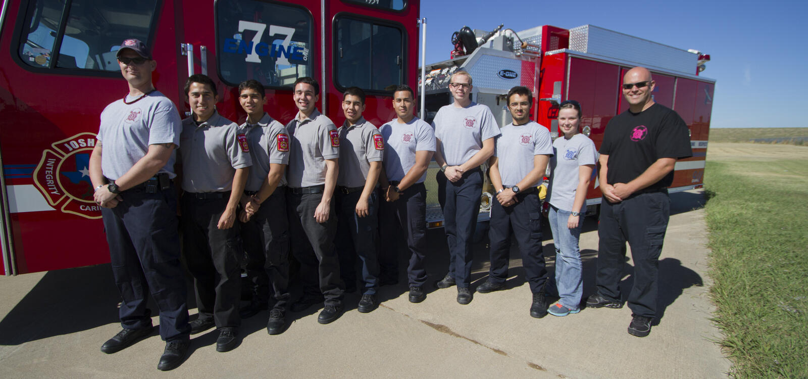 A group of students, matching in gray shirts and dark colored pants, smile as they stand in front of a firetruck