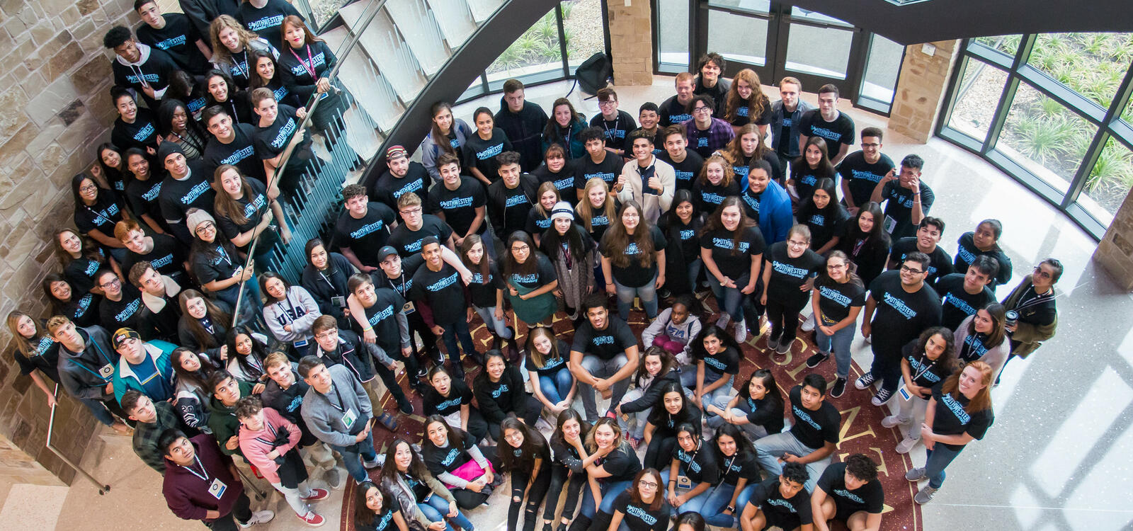 A group of students, all matching in black shirts that say "Southwestern Adventist University" in blue letters, look up as the stand on stairs and on the floor, smiling and looking up