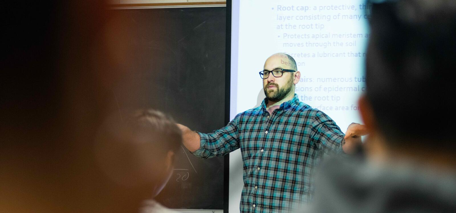 A biology professor stands in front of his projected slides and speaks to his class