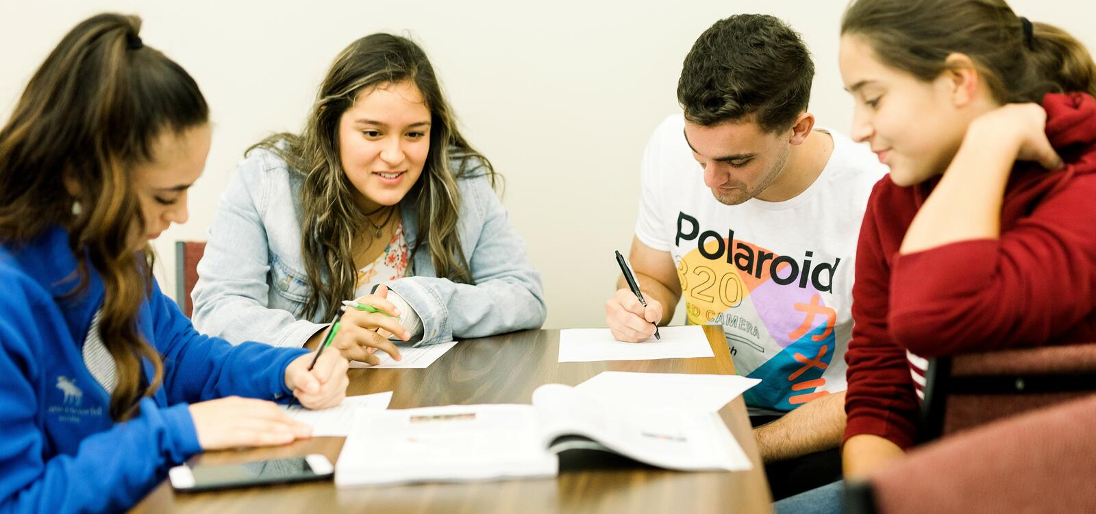 A group of four students sit at a rectangular table as they take notes from a textbook