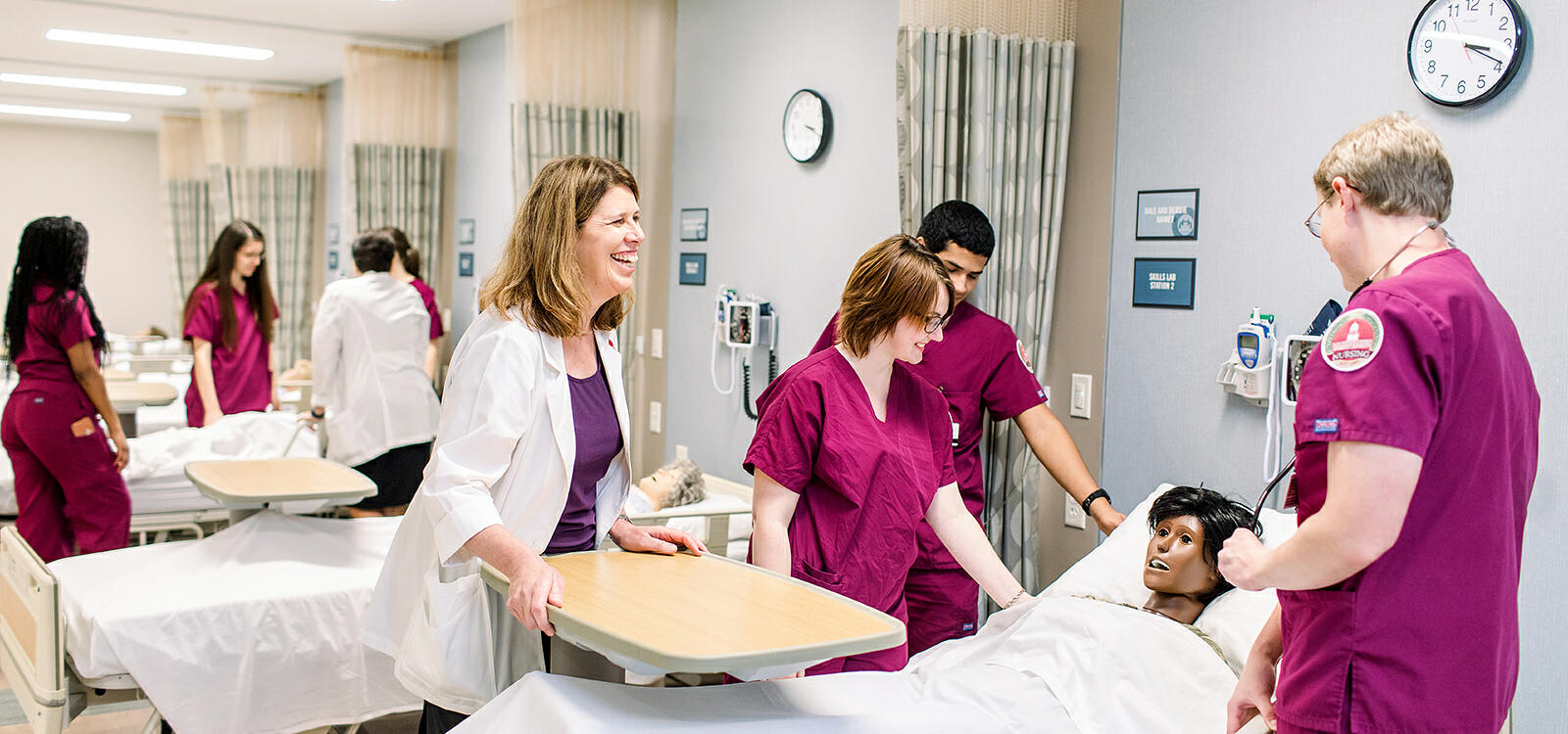 Three nursing students dressed in maroon scrubs look down at their patient and their professor watches over and smiles