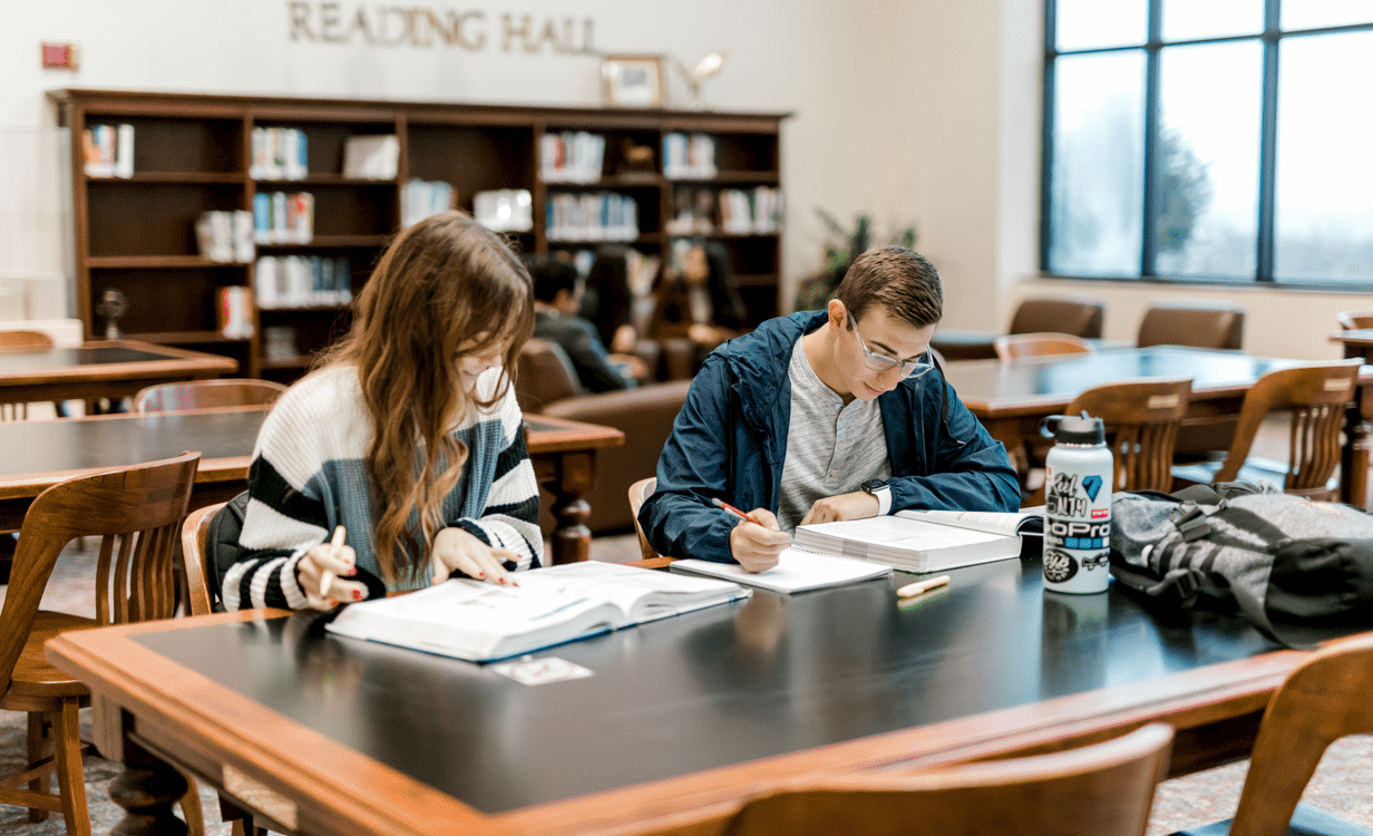 Two students sit at the library table as they read their textbooks and write things down in their notebooks