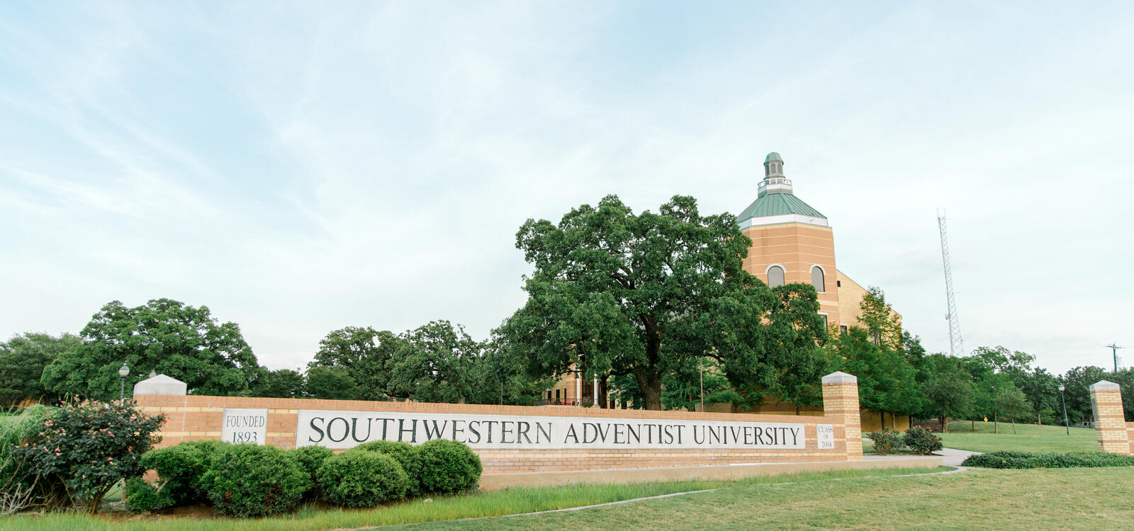 Campus sign made of bricks that reads "Southwestern Adventist University" with the top of Pechero Hall showing behind trees