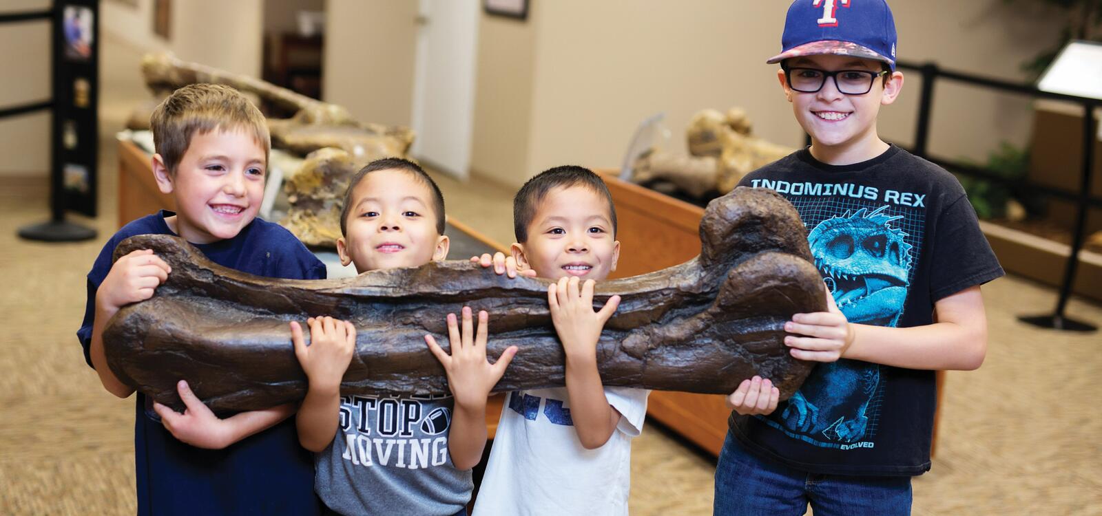 Four young boys smile big as they successfully carry a dinosaur bone that is much bigger than them