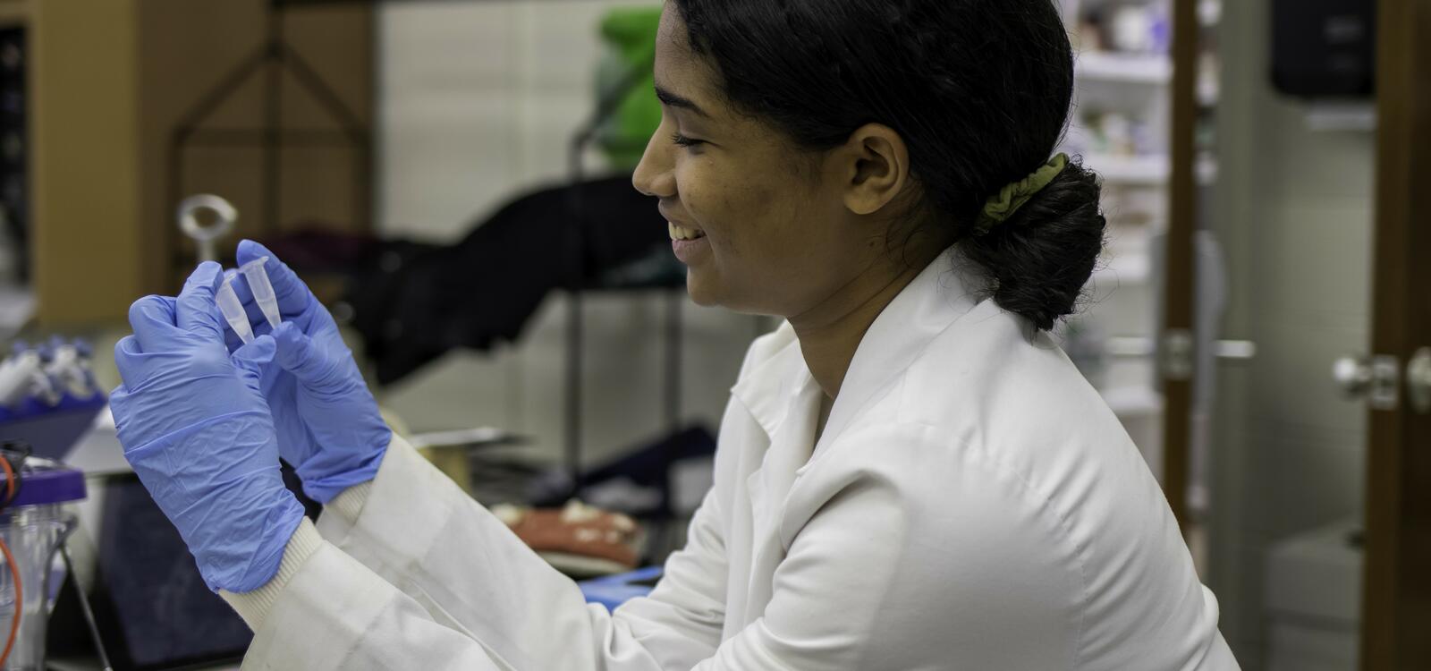 While wearing blue gloves and a lab coat, a student smiles as she picks up two lab tools and observes them