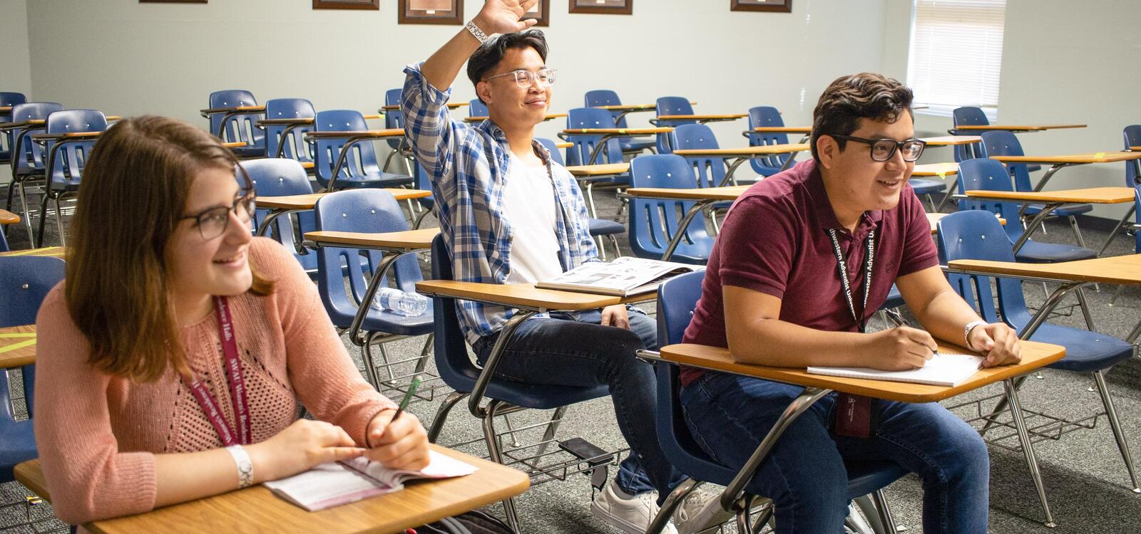 Three students sitting in a classroom facing the front as the listen attentively and take notes. One student is raising his hand to ask a question. 