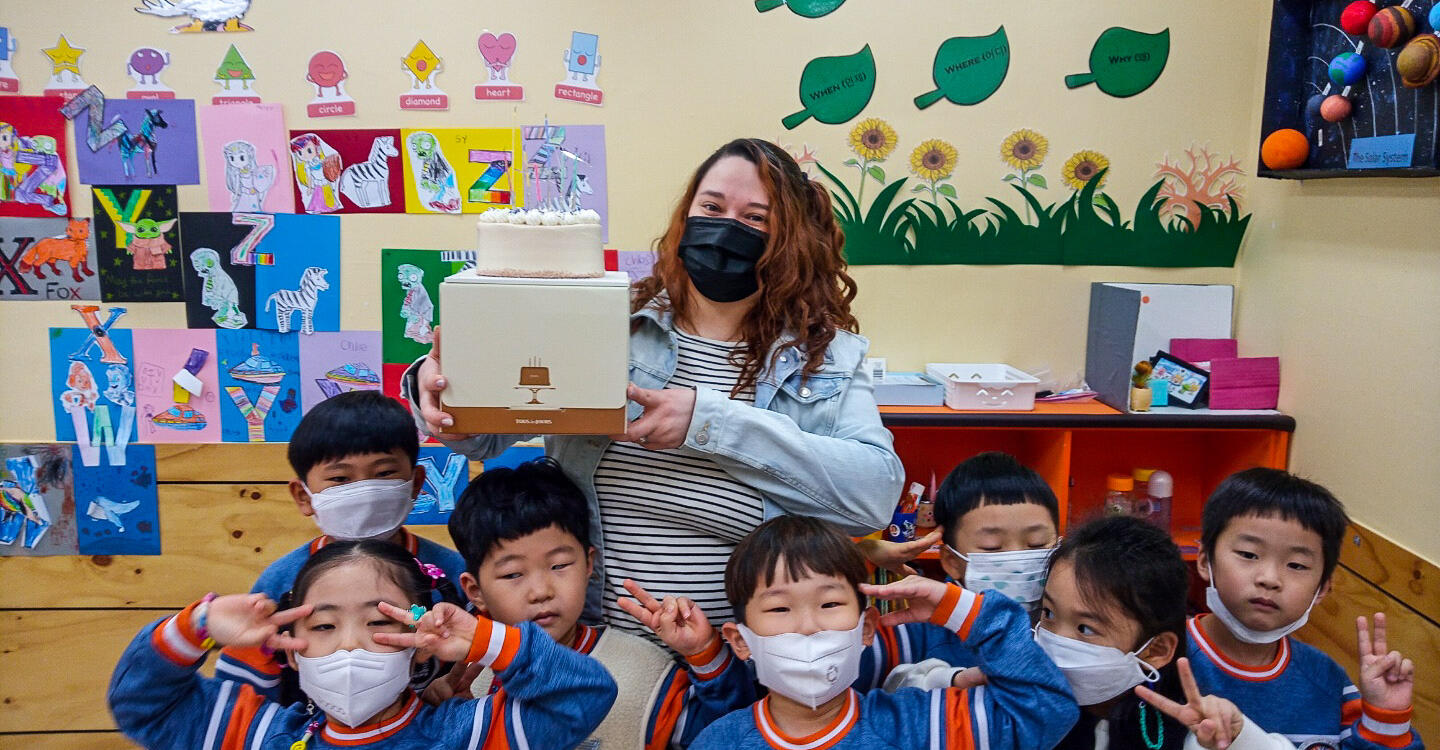 Deanna holds up a box with a cake on top in a group of young students in a classroom
