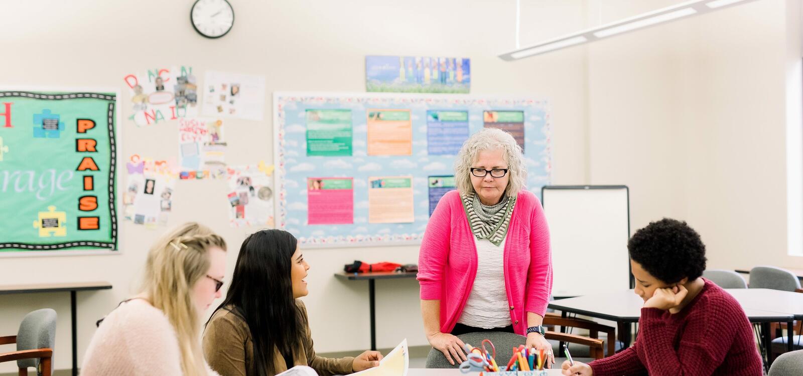 In an educational classroom filled with colorful posters, a professor stands beside a rounded desk, speaking to her students