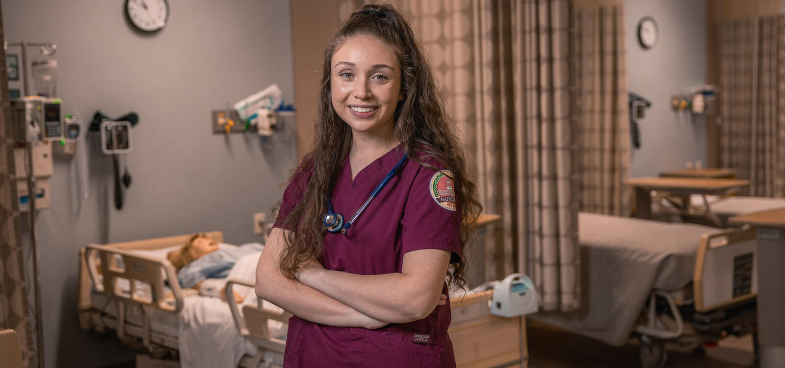 Heather Chirinos poses in the Nursing SIM Lab with a mannequin behind her wearing SWAU scrubs and a stethoscope