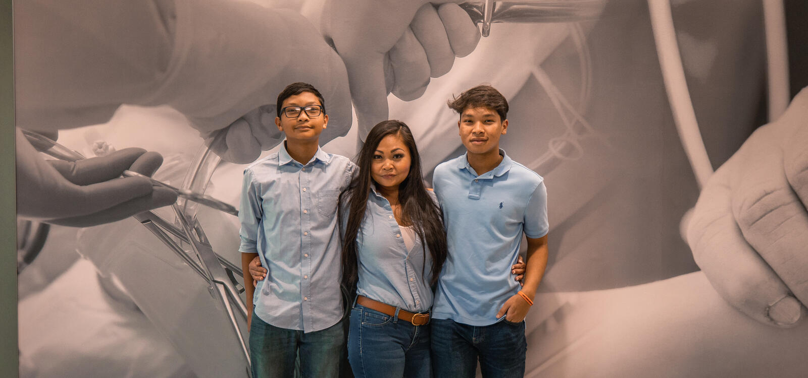 Karen Phongsavan and her two sons pose in front of a wall in the Southwestern Adventist University nursing building that says "Nursing reflects the character of Jesus"