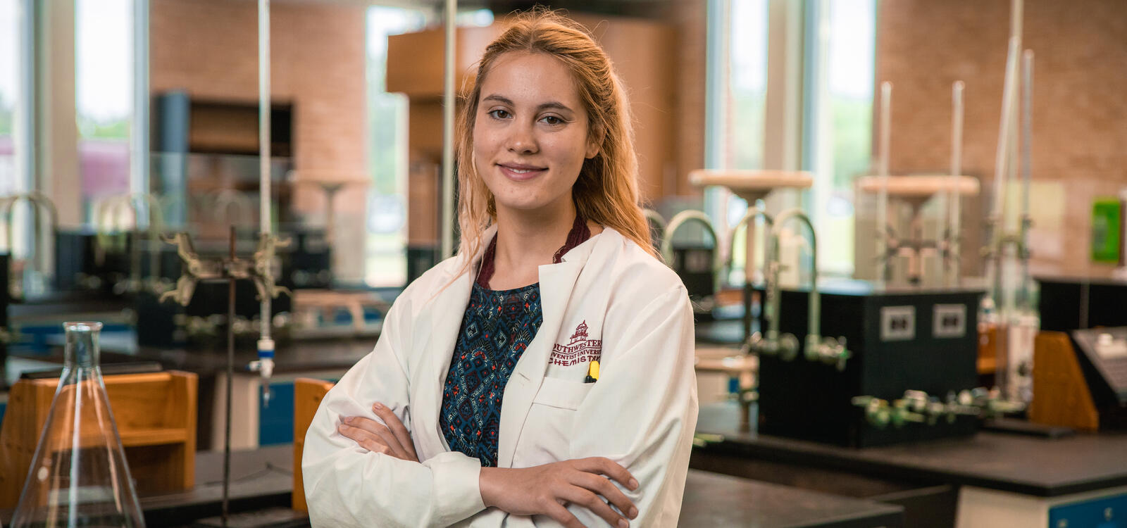 Kelsey Johnson poses in the lab with her arms crossed wearing a lab coat that reads "Southwestern Adventist University Chemistry"