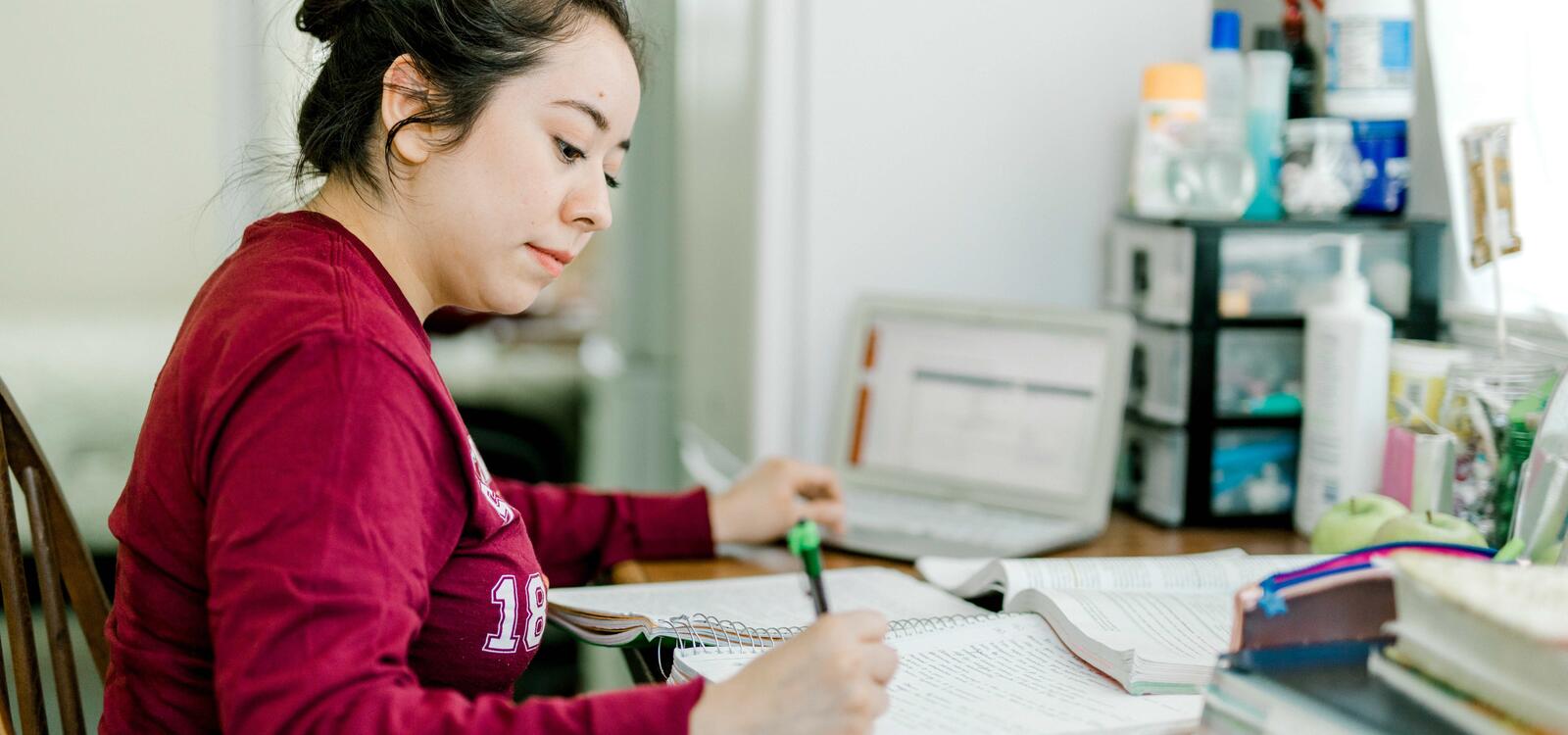 A student, dressed in a maroon long-sleeved shirt and with her hair in a bun, sits in front of a table as she completes her homework 