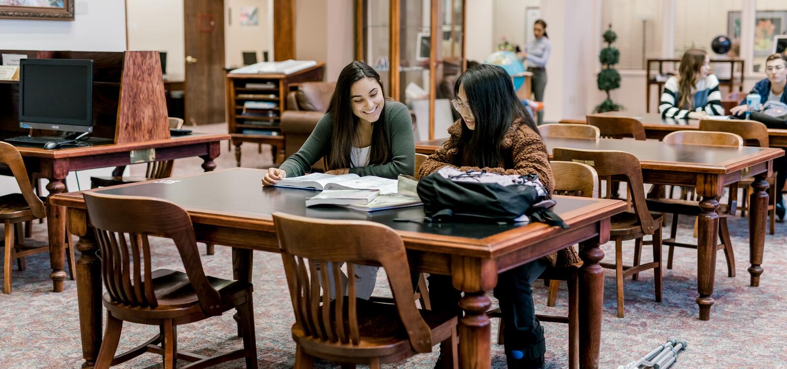 Two female students smiling as the sit at a table at the library and read through a textbook